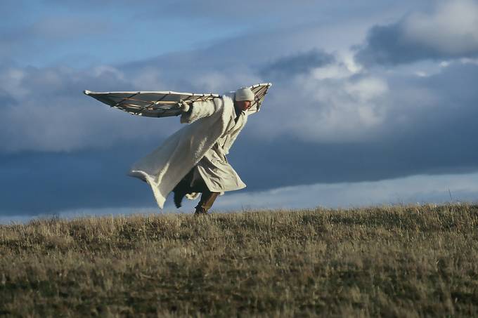 Weiß gekleideter Mann mit gebastelten Flügeln rennt einen Hügel hinauf, Wolken im Hintergrund