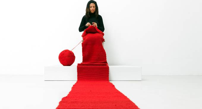 Eine Frau sitzt in einem weißen Raum auf einem Podest und strickt an einem langen roten Schal