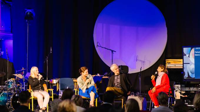 Auf einer Bühne sitzen drei Frauen und ein Mann. Die Moderatorin trägt einen roten Hosenanzug. Im Hintergrund eine schwarze Fläche mit einer großen Runden Fläche, die an einen Mond erinnert  (öffnet Vergrößerung des Bildes)