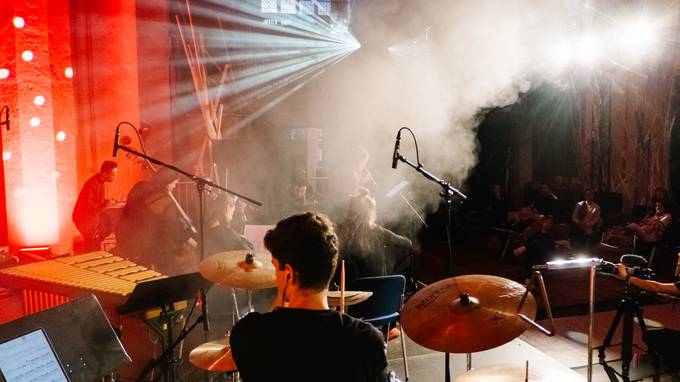 Ein Schlagzeuger auf einer Bühne fotografiert von hinten, mehrere Mikrofone, besondere Lichtspiele mit Nebel (öffnet Vergrößerung des Bildes)