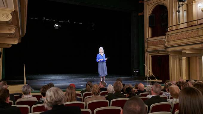 Eine Frau in blauer Abendgarderobe steht auf einer Theaterbühne, im Bildvordergrund sind mehrere Publikumsreihen zu sehen. (öffnet Vergrößerung des Bildes)