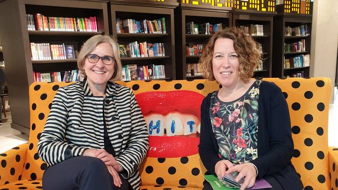 Zwei Frauen auf Sofa lächeln in die Kamera, im Hintergrund Bücherregale (öffnet Vergrößerung des Bildes)