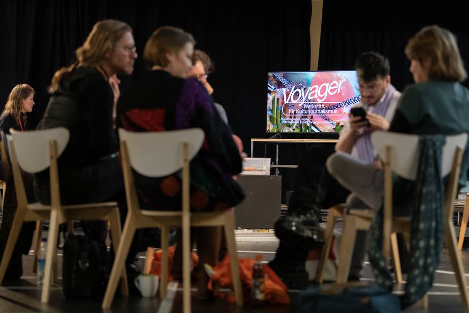 Eine Gruppe von ca. 20 Menschen in einem Bühnenraum. Sie sitzen im Stuhlkreis und kleinen Gruppen zusammen, diskutieren, schauen auf Tabletcomputer und sammeln Stichworte an einem Flipchart.