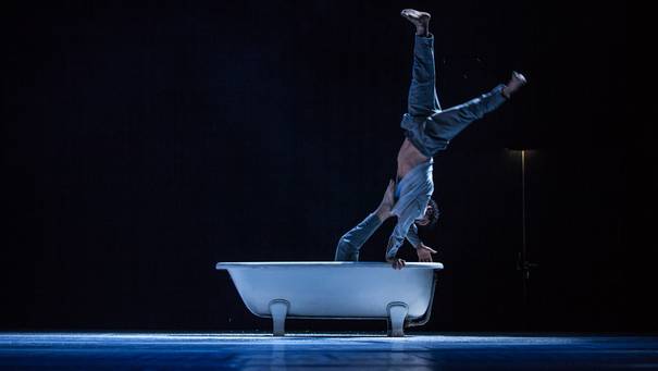 Foto einer Badewanne auf dunkel ausgeleuchteter Bühne, auf deren Rand eine Tänzerin Handstand macht