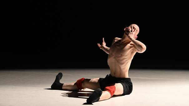 Ein Tänzer mit schwarzer Hose, Schienbeinschonern und nacktem Oberkörper sitzt seitlich angewinkelt mit ausgestreckten Beinen auf dem Boden und hält eine Hand vor sein Gesicht