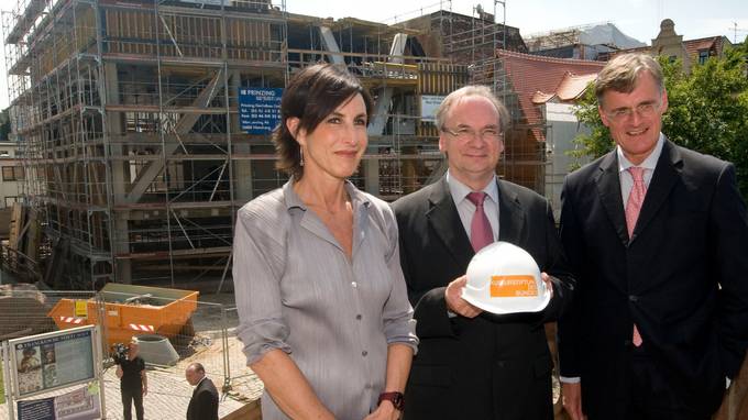 Eine Frau und zwei Männer, einer davon mit Helm in der Hand, posieren vor dem Rohbau eines Gebäudes (öffnet Vergrößerung des Bildes)