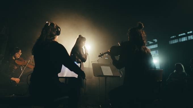 Auf der Bühne steht eine Sängerin in einem Lichtkegel, umgeben von spielenden Streicherinnen. (öffnet Vergrößerung des Bildes)
