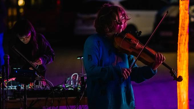 Es ist dunkel. Man erkennt eine Frau, die Geige spielt, und im Hintergrund eine weitere Frau an einem Mischpult (öffnet Vergrößerung des Bildes)