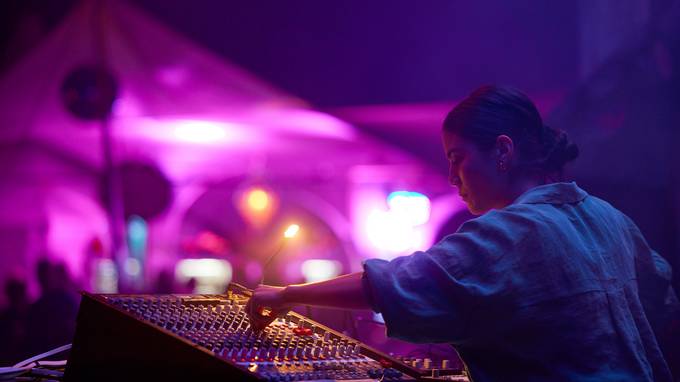 Eine Frau steht an einem DJ-Mischpult. Es ist dunkel, im Hintergrund leuchtet ein Festivalzelt in pinkem Licht. (öffnet Vergrößerung des Bildes)